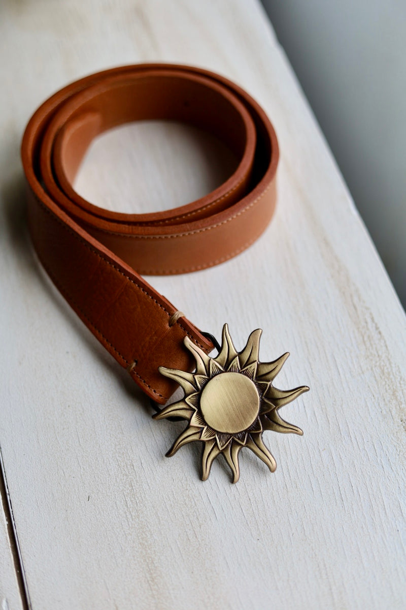 Brown leather belt with a sun-shaped brass buckle on a light wooden surface.