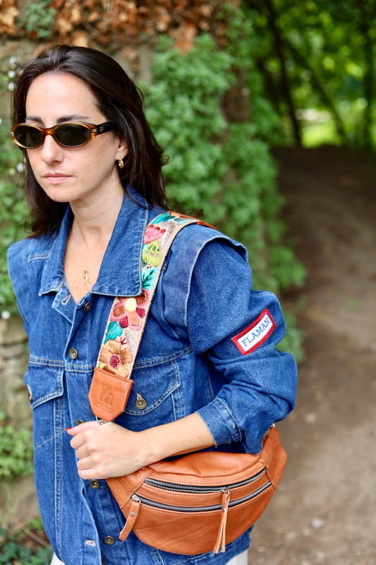 A woman wearing a blue denim jacket and sunglasses carries a tan leather fanny pack with a colorful floral strap and a red-and-white patch on the sleeve, standing outdoors on a path with green foliage in the background.