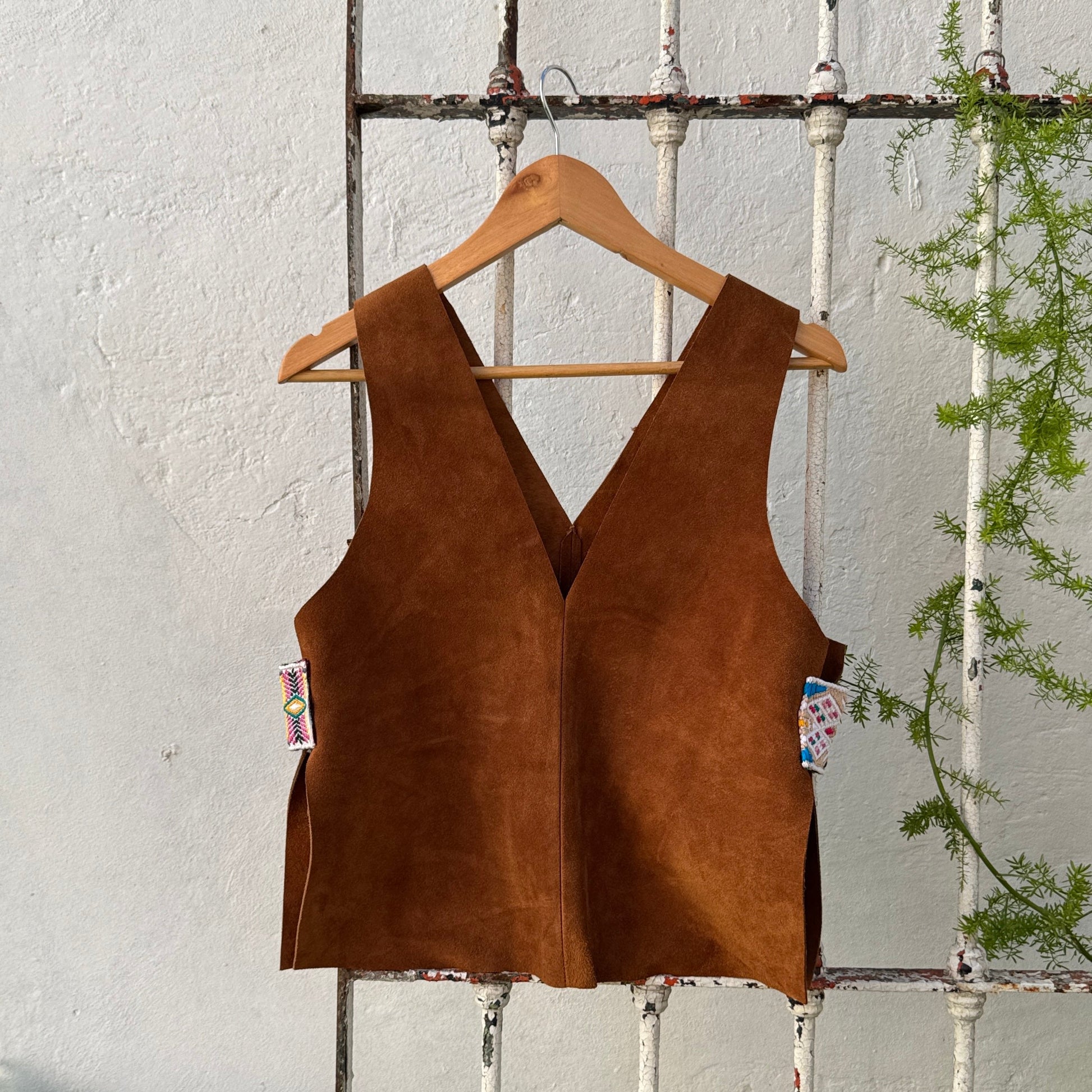 Brown suede vest on a hanger against a rustic white wall with greenery.