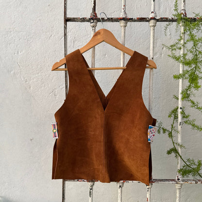 Brown suede vest on a hanger against a rustic white wall with greenery.