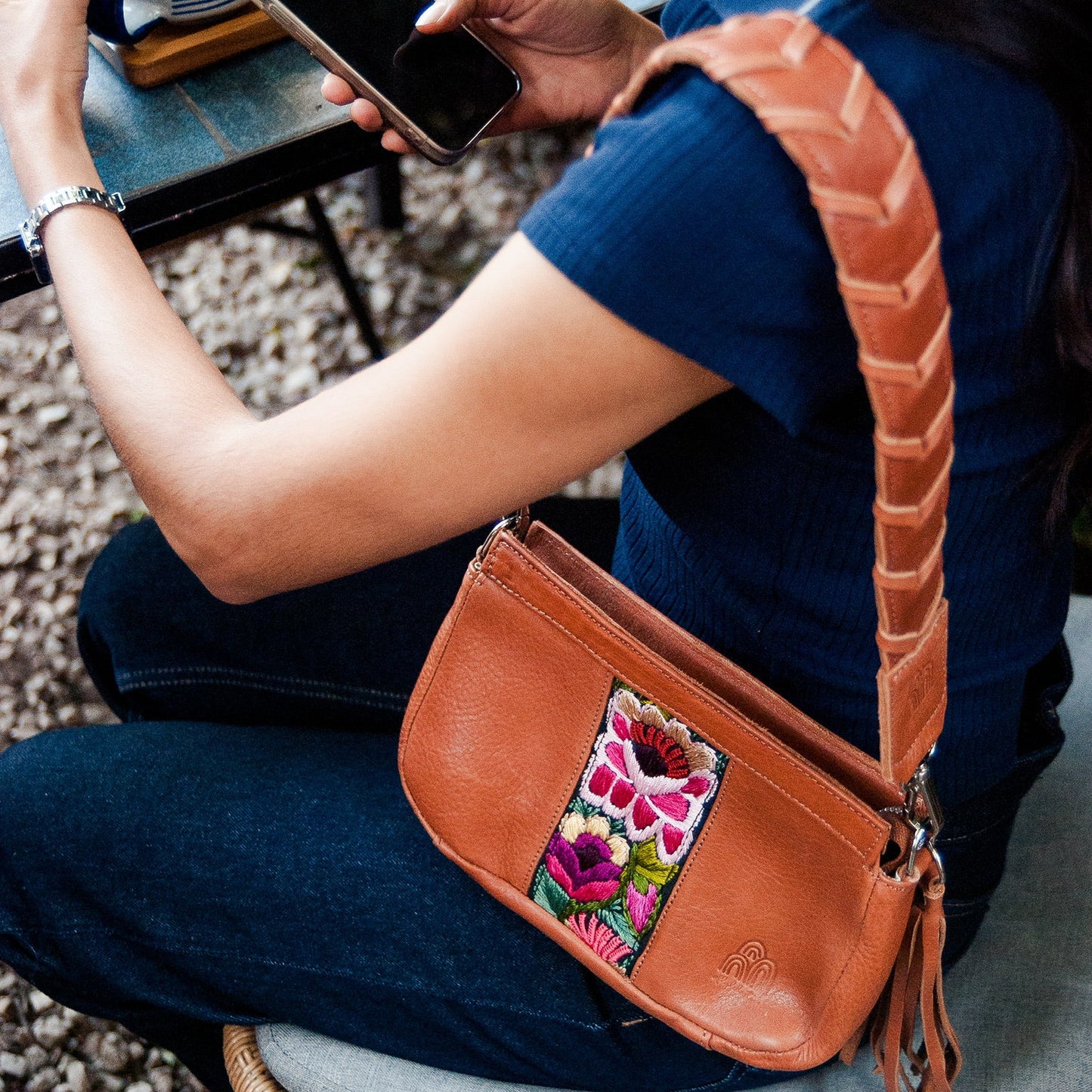 Person sitting on a bench using a smartphone with a brown handbag featuring floral embroidery.