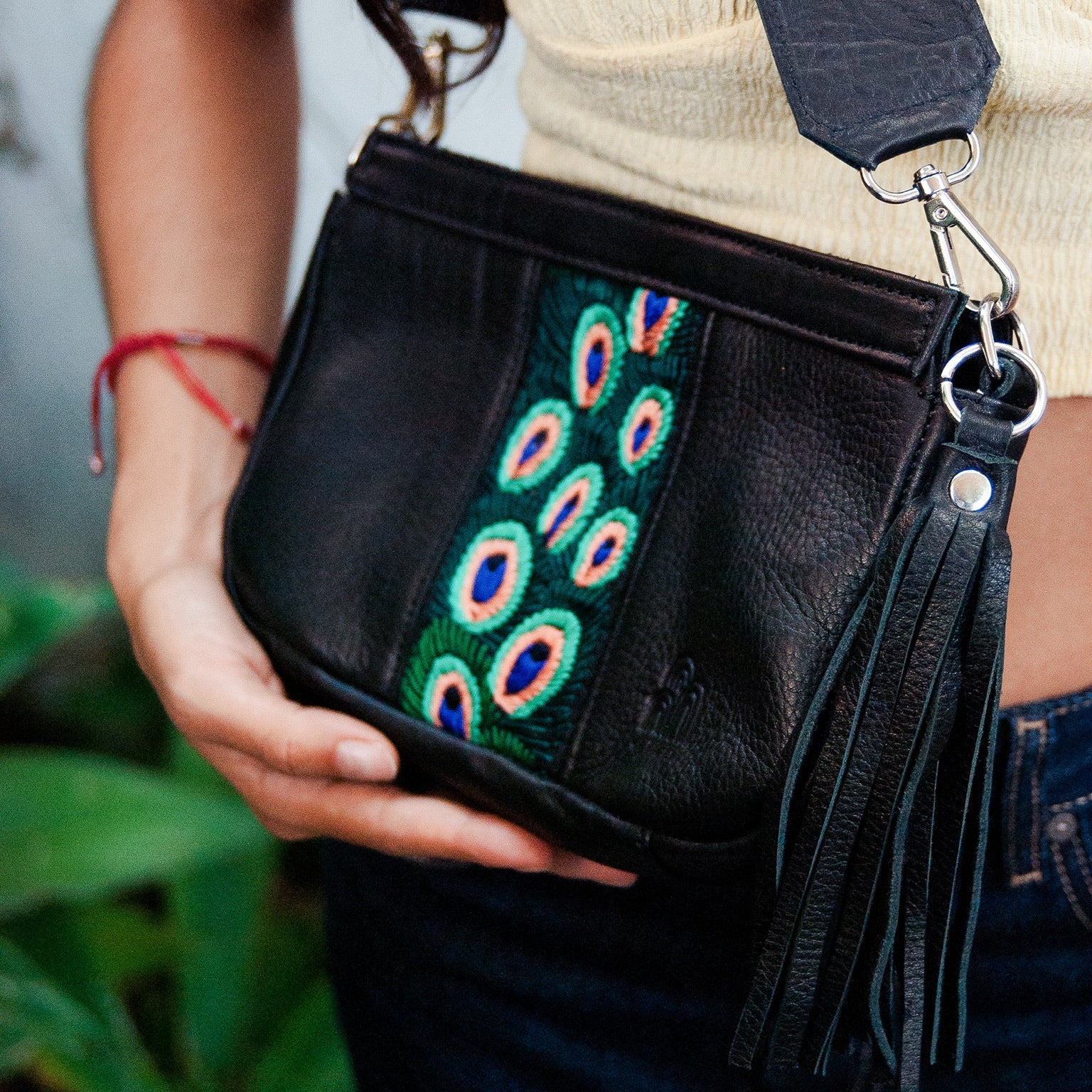 Black handbag with peacock feather strap held by a person, blurred green plants in the background