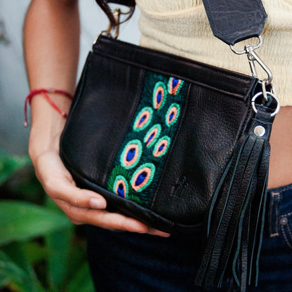 Black handbag with peacock feather strap held by a person, blurred green plants in the background