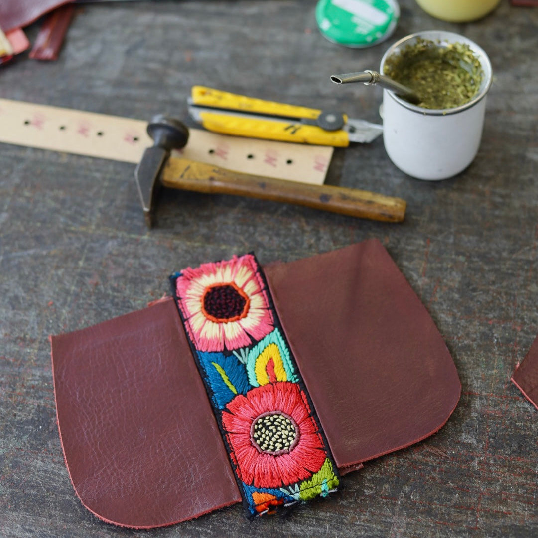 Colorful floral embroidered leather strap on a dark surface with tools and a mug.
