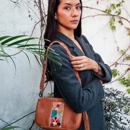 Woman holding a brown leather bag with floral patterns against a white wall with greenery.