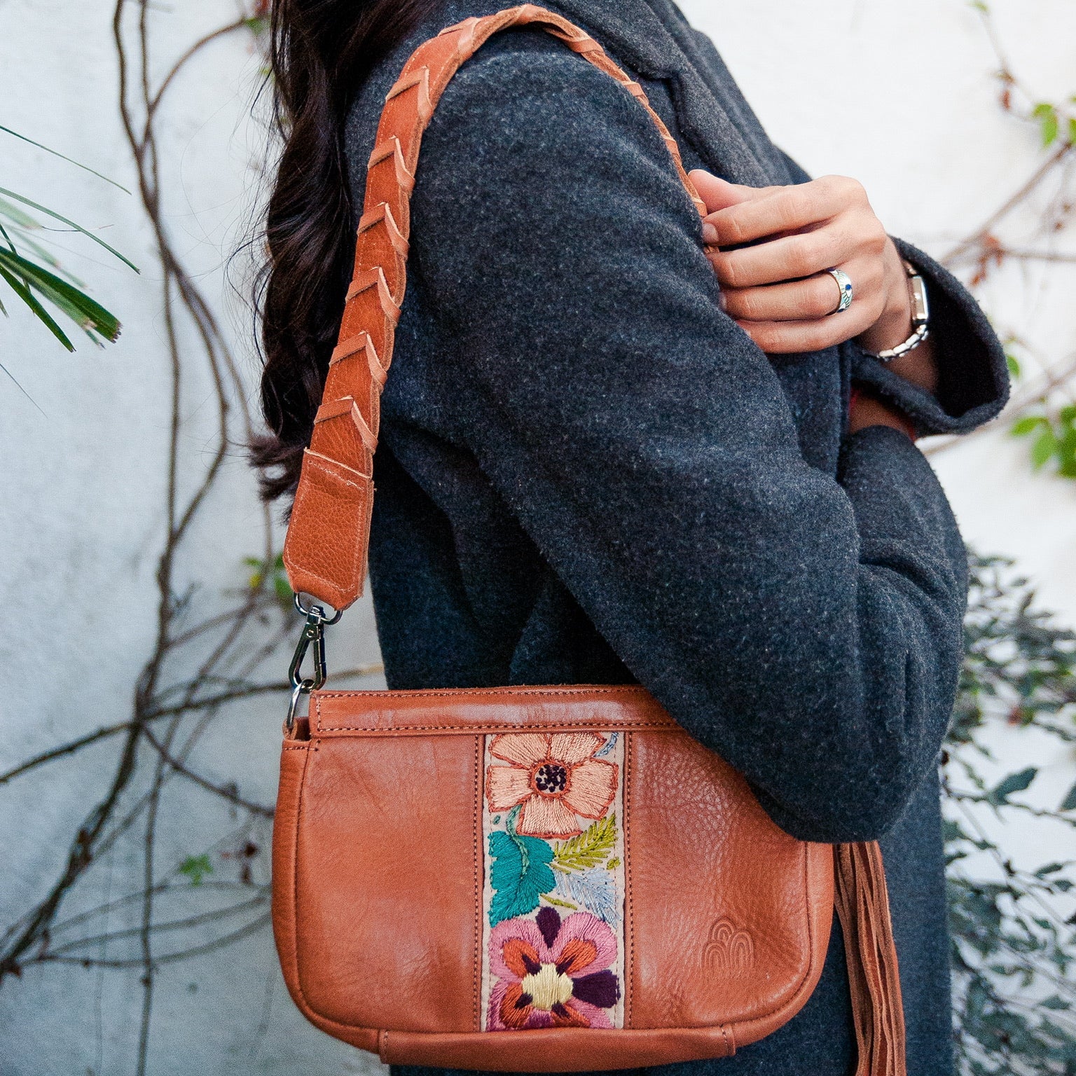 Person holding a brown leather handbag with floral patchwork design against a white wall.