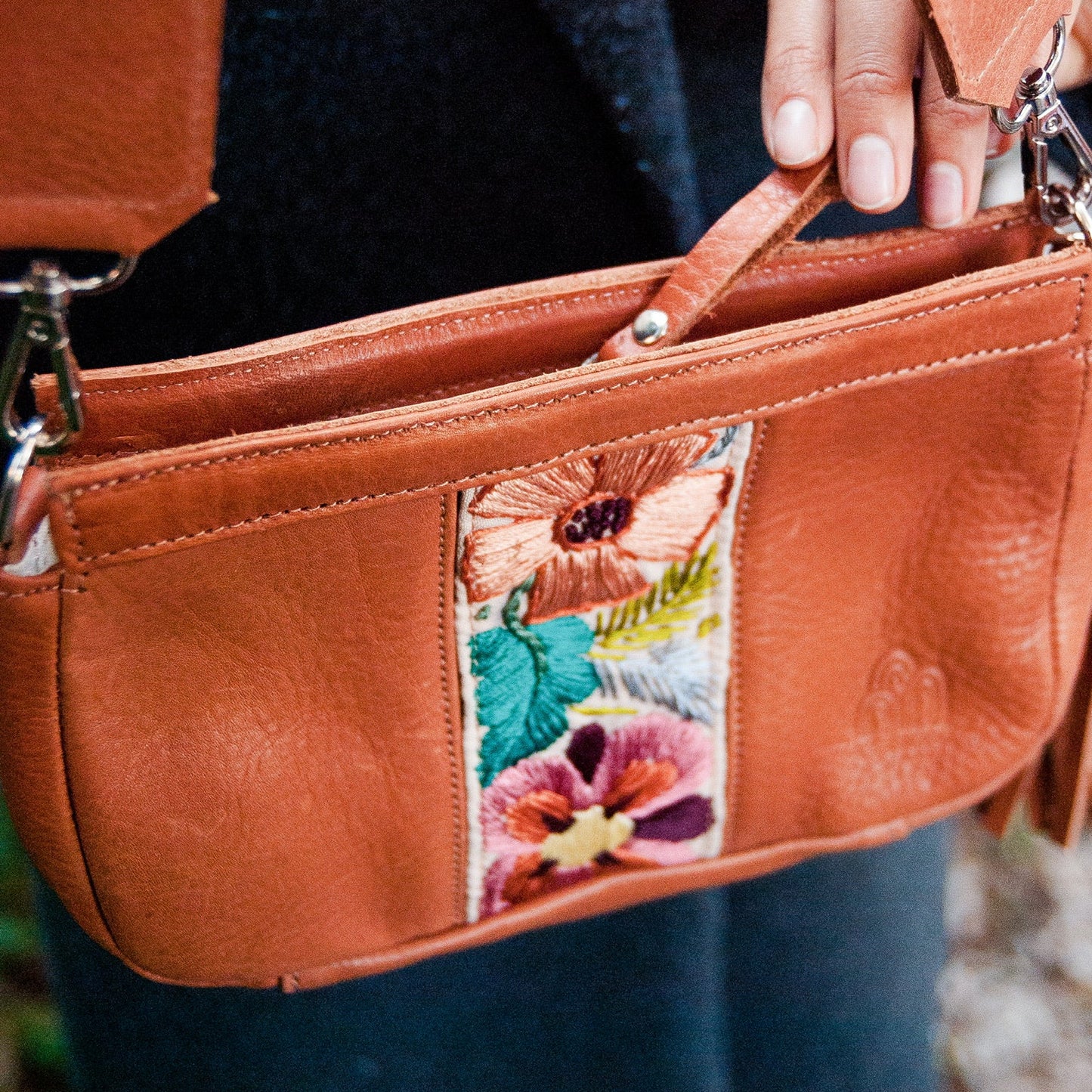 Brown leather handbag with floral patch held by a person wearing a dark coat.