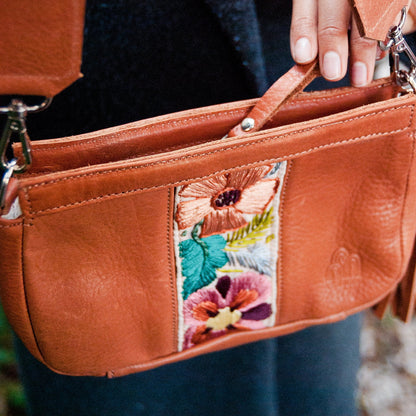 Brown leather handbag with floral patch held by a person wearing a dark coat.