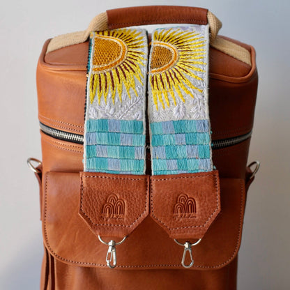 A brown leather picnic bag with a pair of hand-knotted straps featuring a geometric pattern in blue and yellow, and a tan exterior pocket.