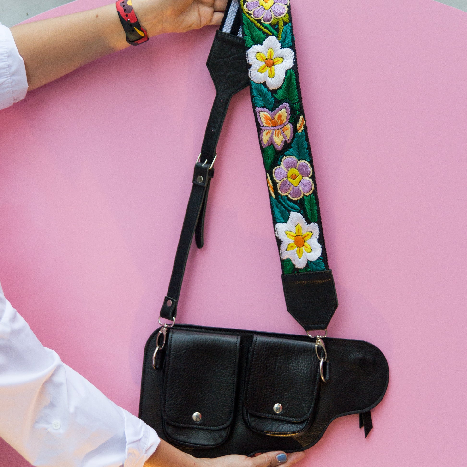 Black bag with a floral strap held by a person on a pink surface