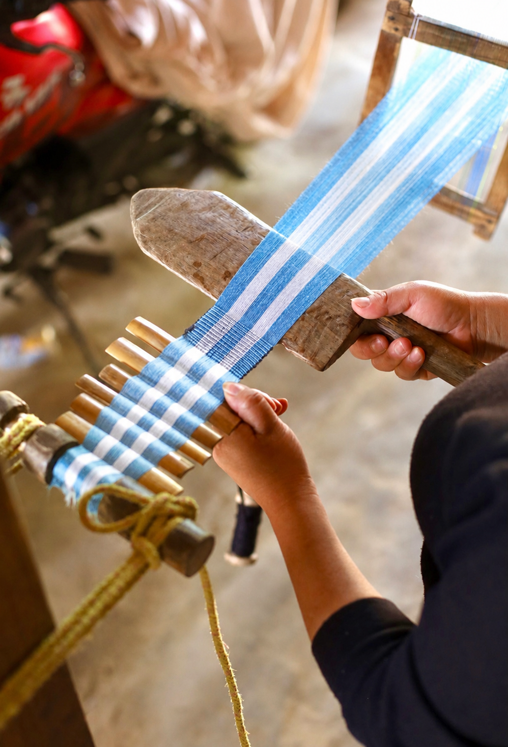 A close-up of a person weaving a blue and white striped fabric using a traditional wooden loom with wooden mallets.