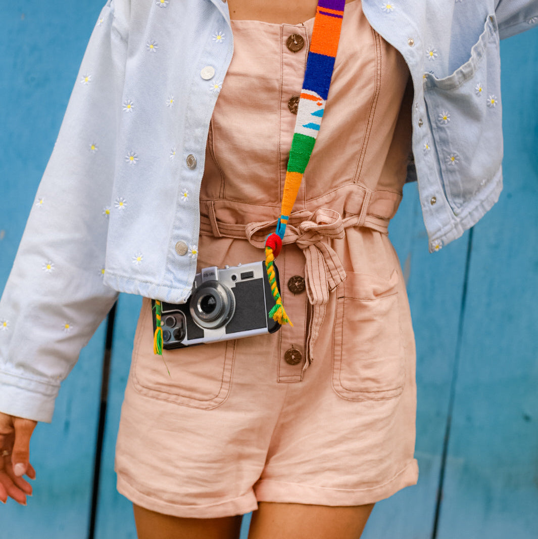 Person wearing a light blue denim jacket over a beige outfit with a colorful belt and camera, against a blue wooden background.