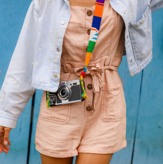 Person wearing a light blue denim jacket over a beige outfit with a colorful belt and camera, against a blue wooden background.