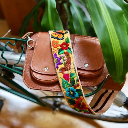 Brown leather bag with a colorful floral strap on a bicycle, surrounded by green plants.