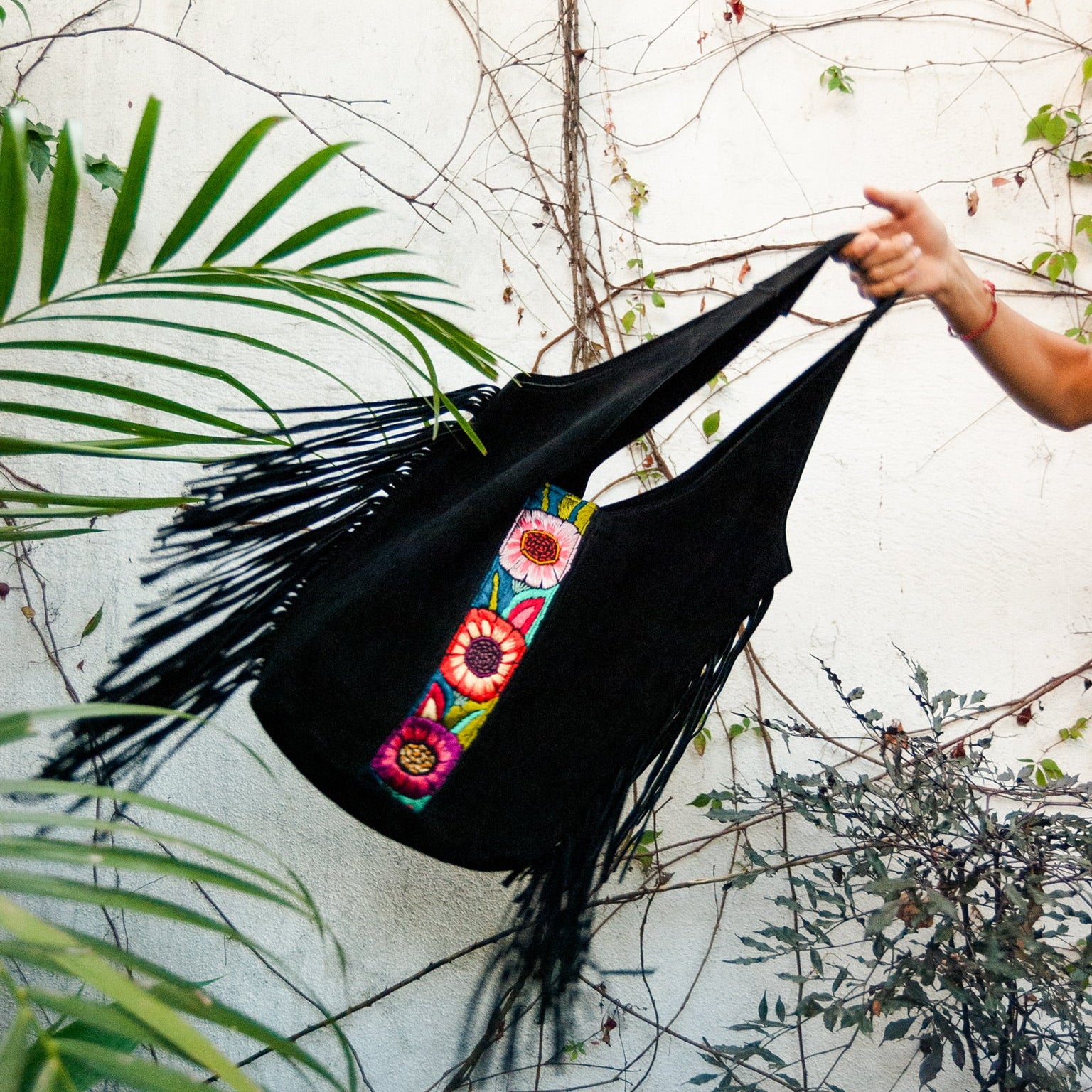 Black hammock with colorful floral embroidery held by a hand against a white wall with greenery.