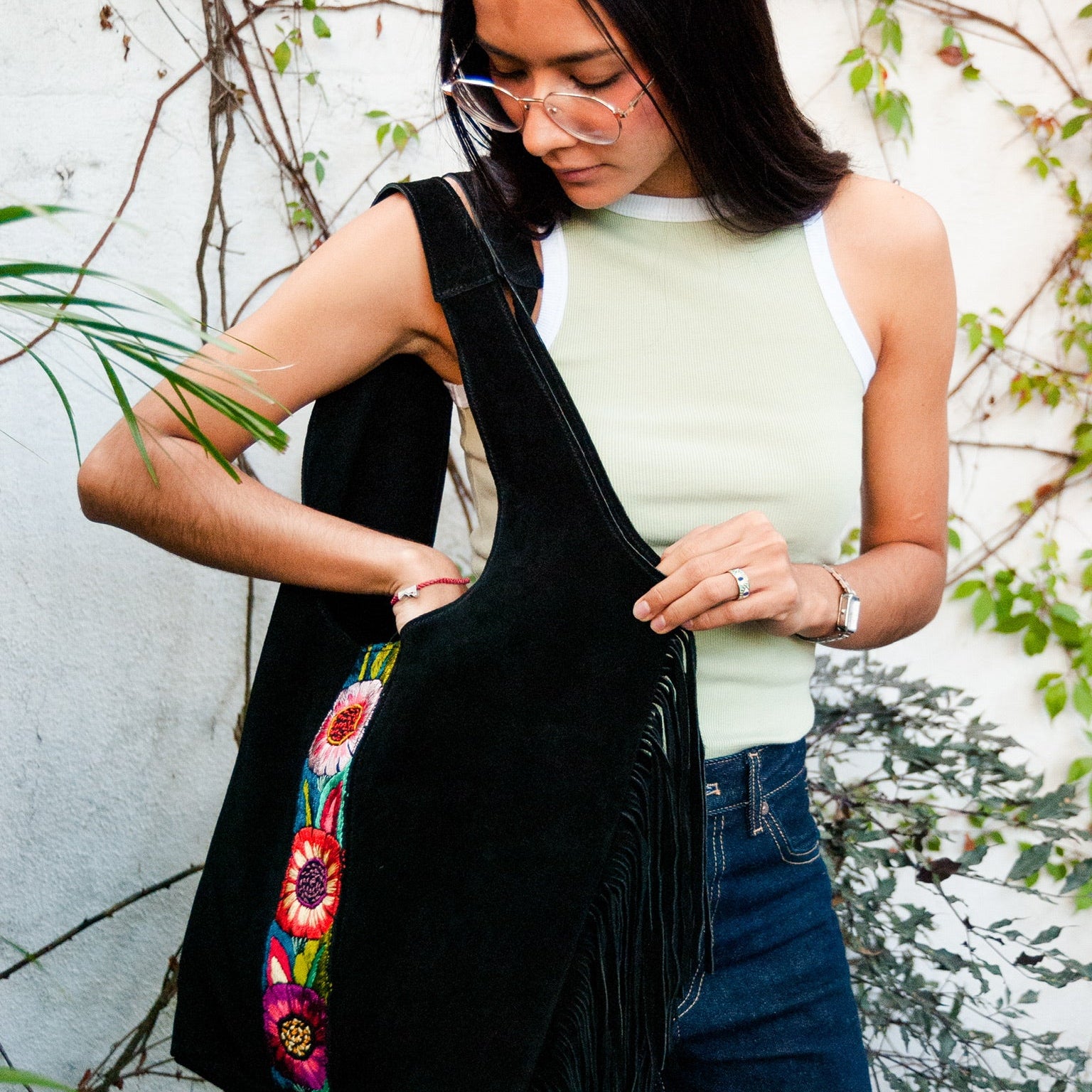 Woman holding a black bag with floral embroidery against a white wall with greenery.