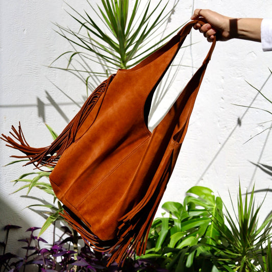 Brown leather bag with fringe held by a hand against a white wall with plants.