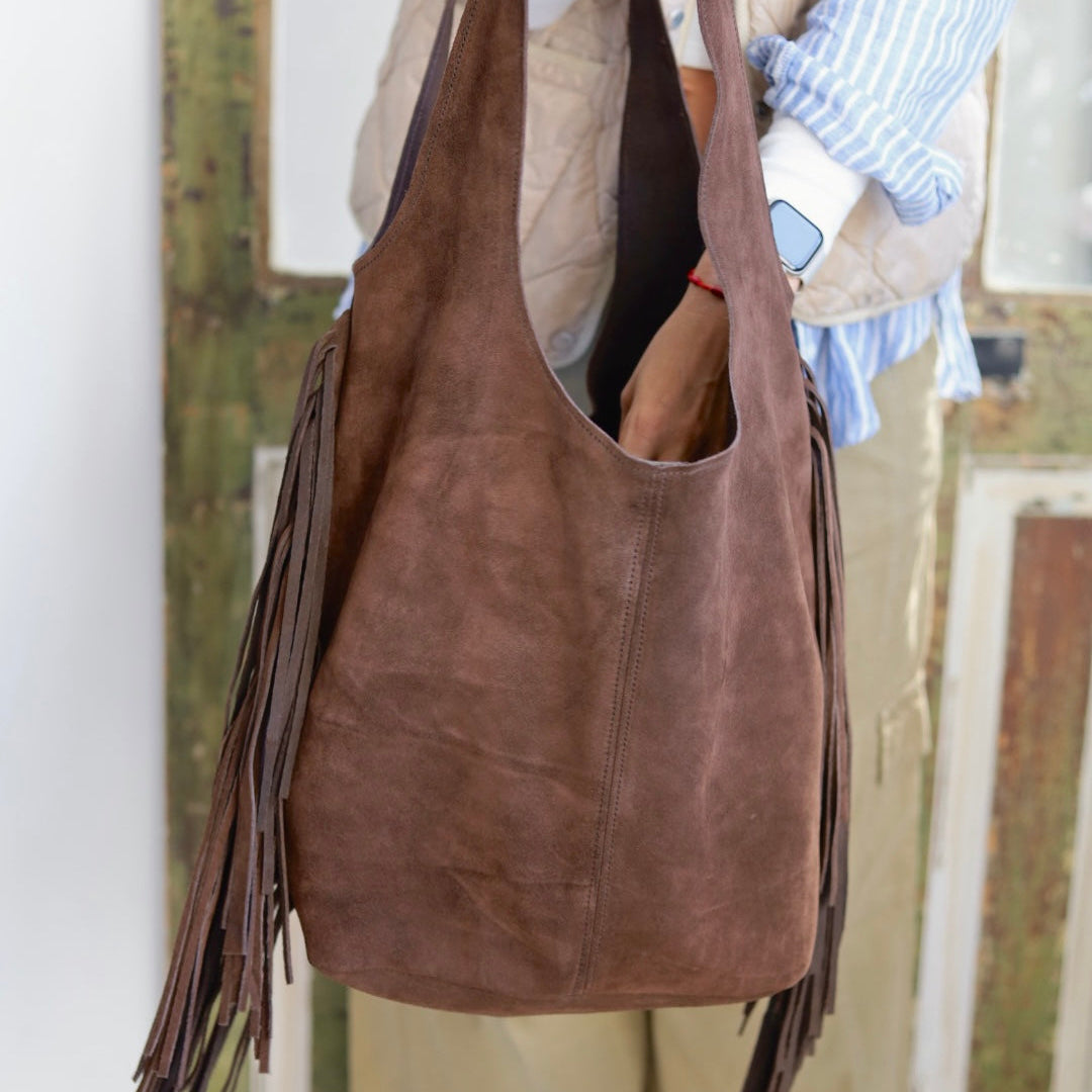 Brown suede handbag with fringe held by a person wearing a blue shirt and beige vest.