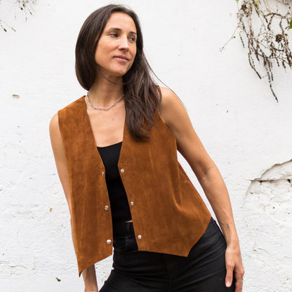 Woman wearing a brown vest over a black top and pants against a white wall with greenery.
