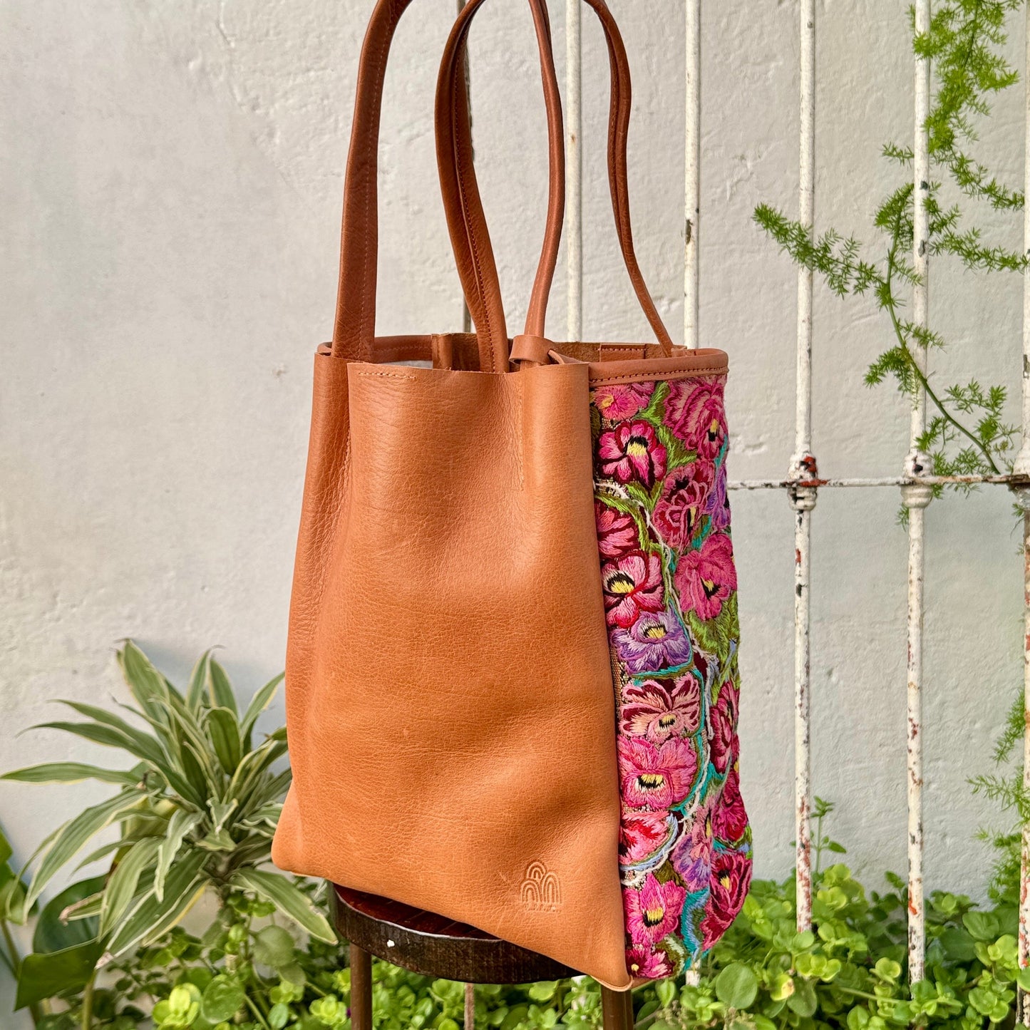 Brown leather tote bag with floral patterned interior on a stand against a white wall with plants.