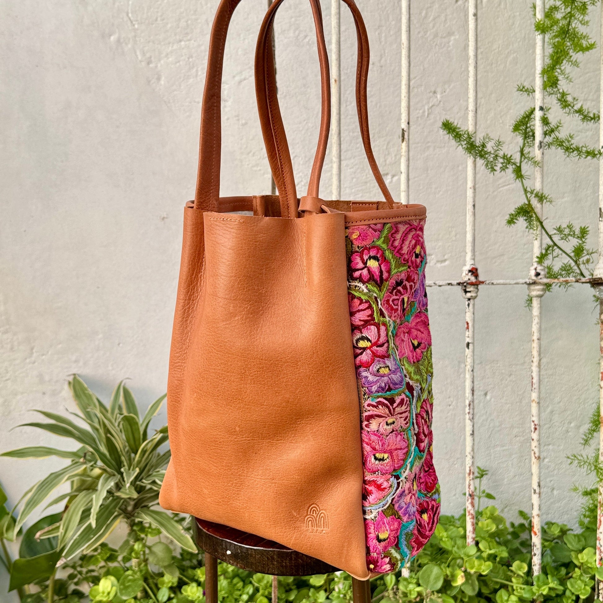 Brown leather tote bag with floral patterned interior on a stand against a white wall with plants.