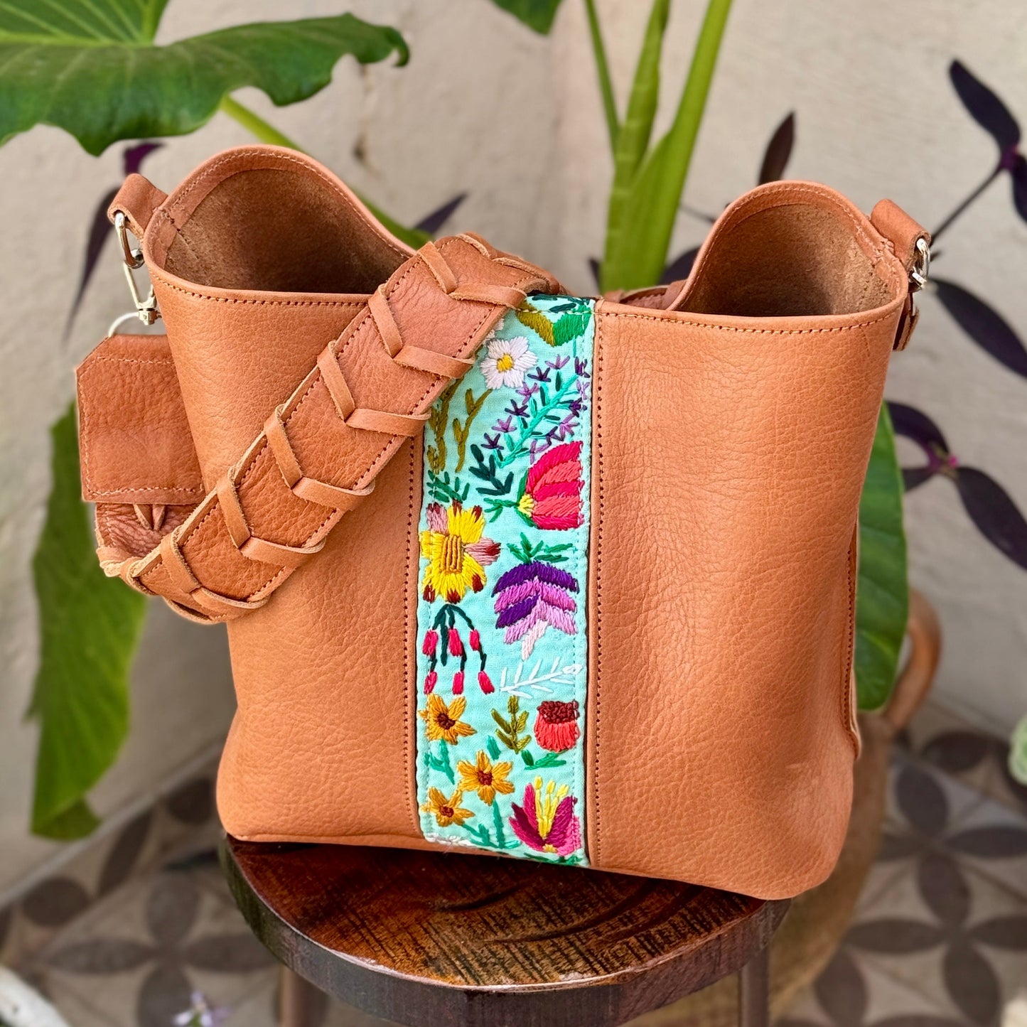 Tan handbag with floral strap on a wooden stool against a patterned wall.