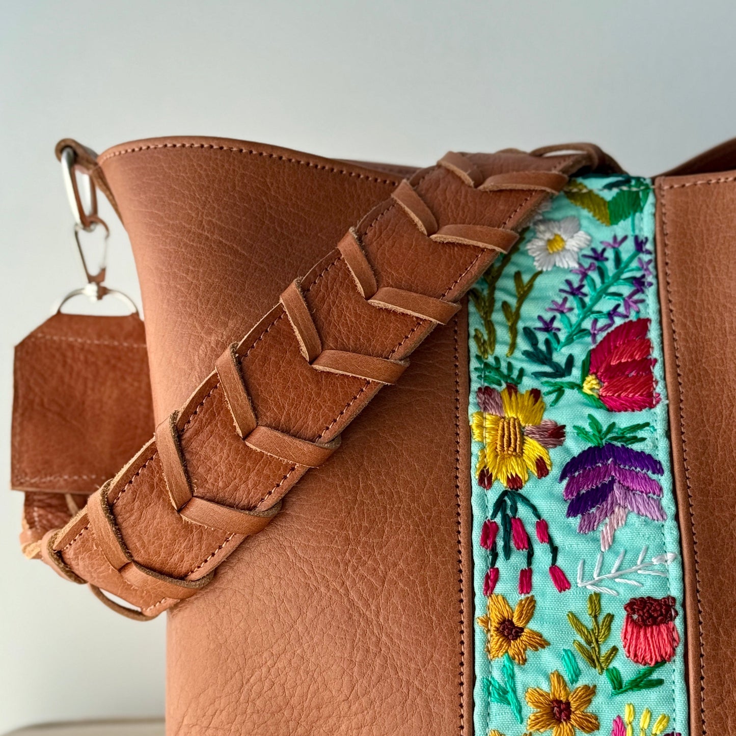 Brown leather handbag with a colorful floral embroidered strap on a light gray background