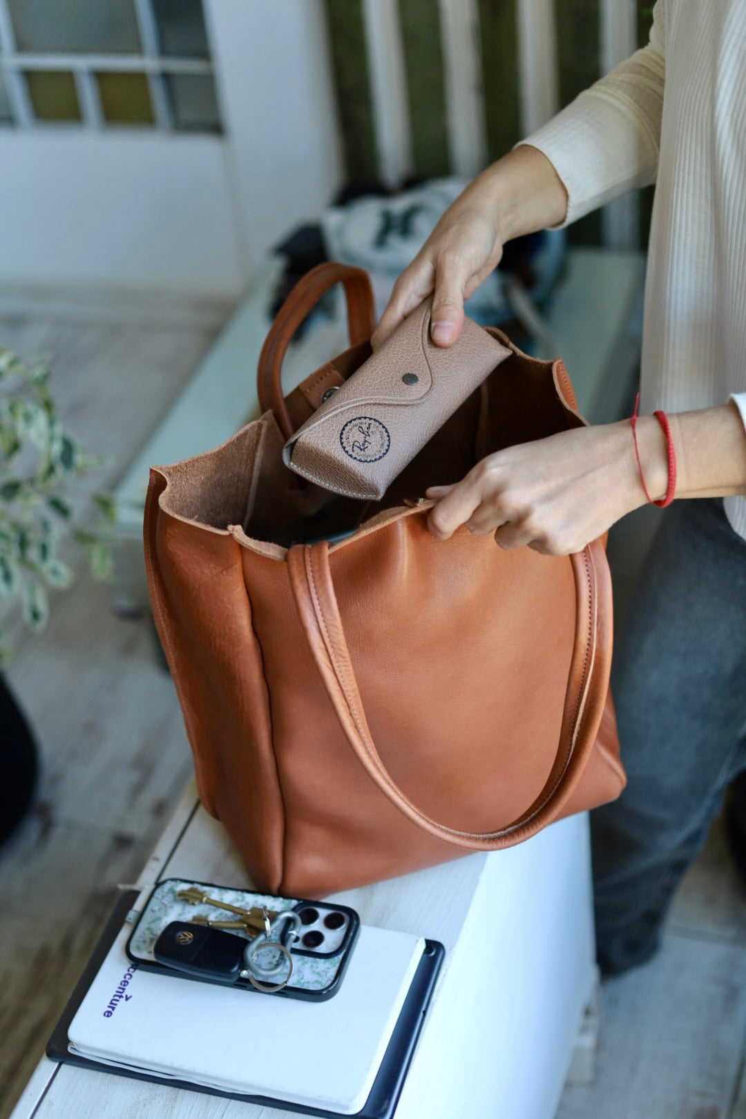 Person opening a brown leather bag with a phone and keys on a surface.