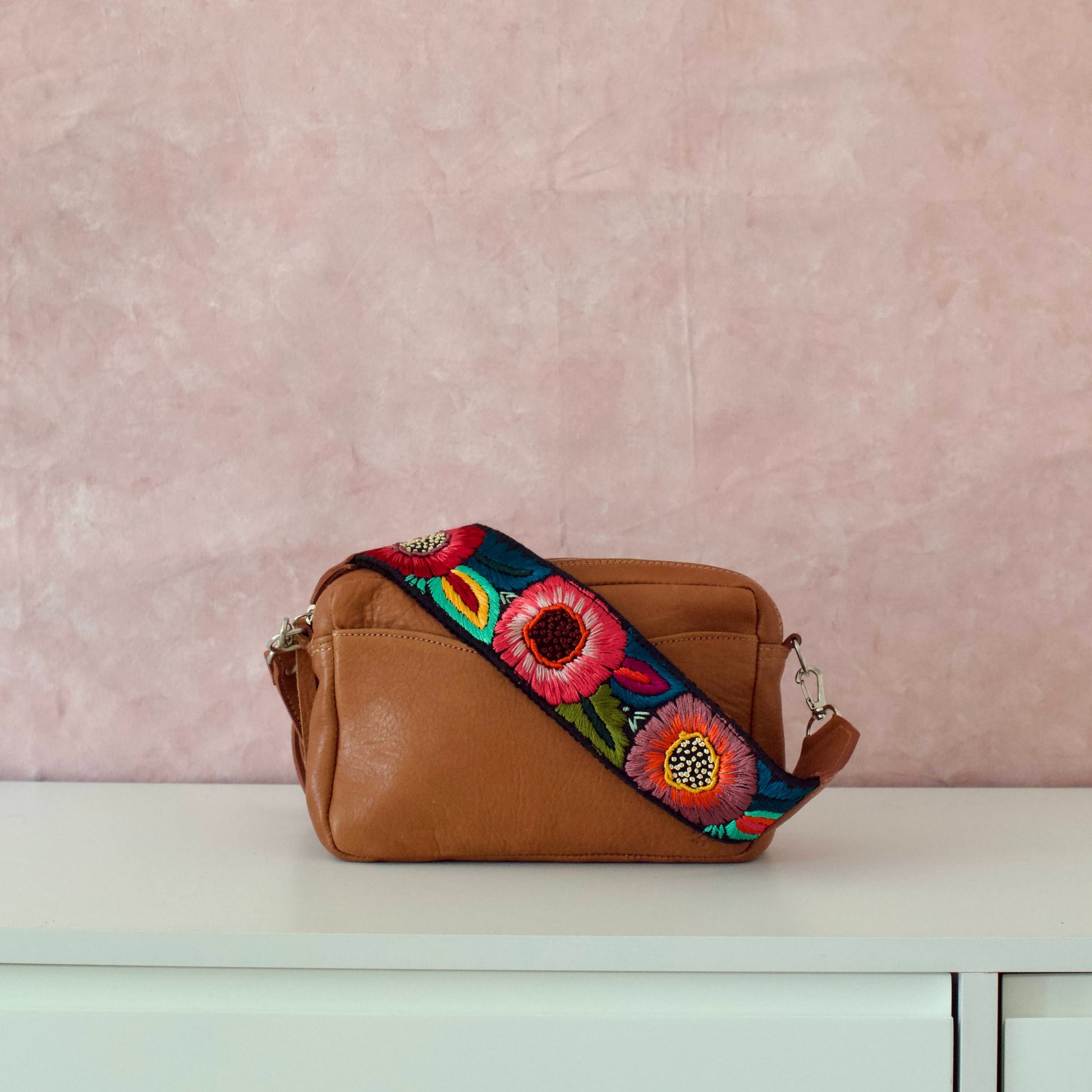 A brown leather travel necessaire Aimé with a multicolored floral strap, displayed against a pink-toned background.