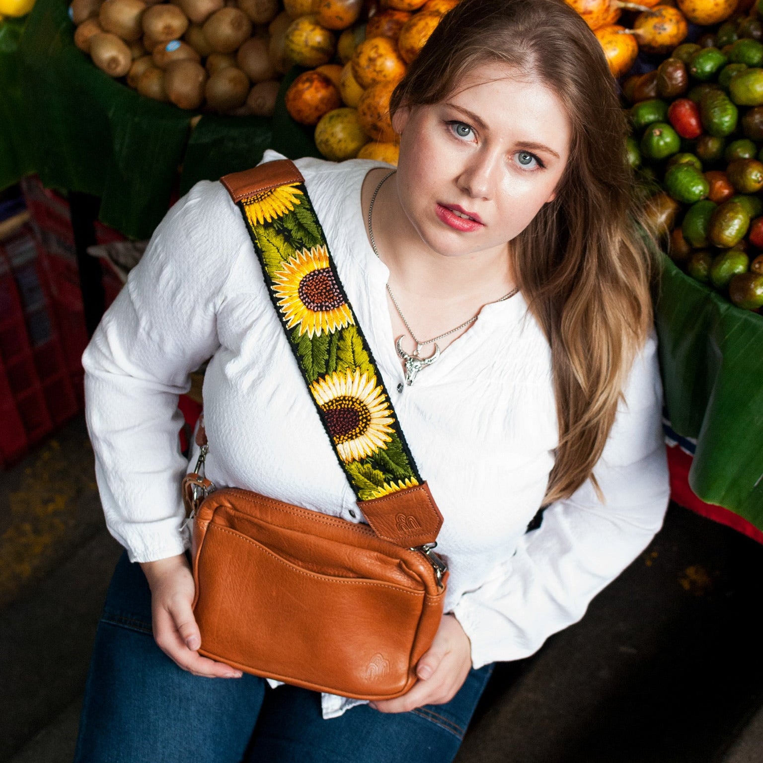 Woman holding a brown leather bag with a colorful strap in front of a fruit stand.