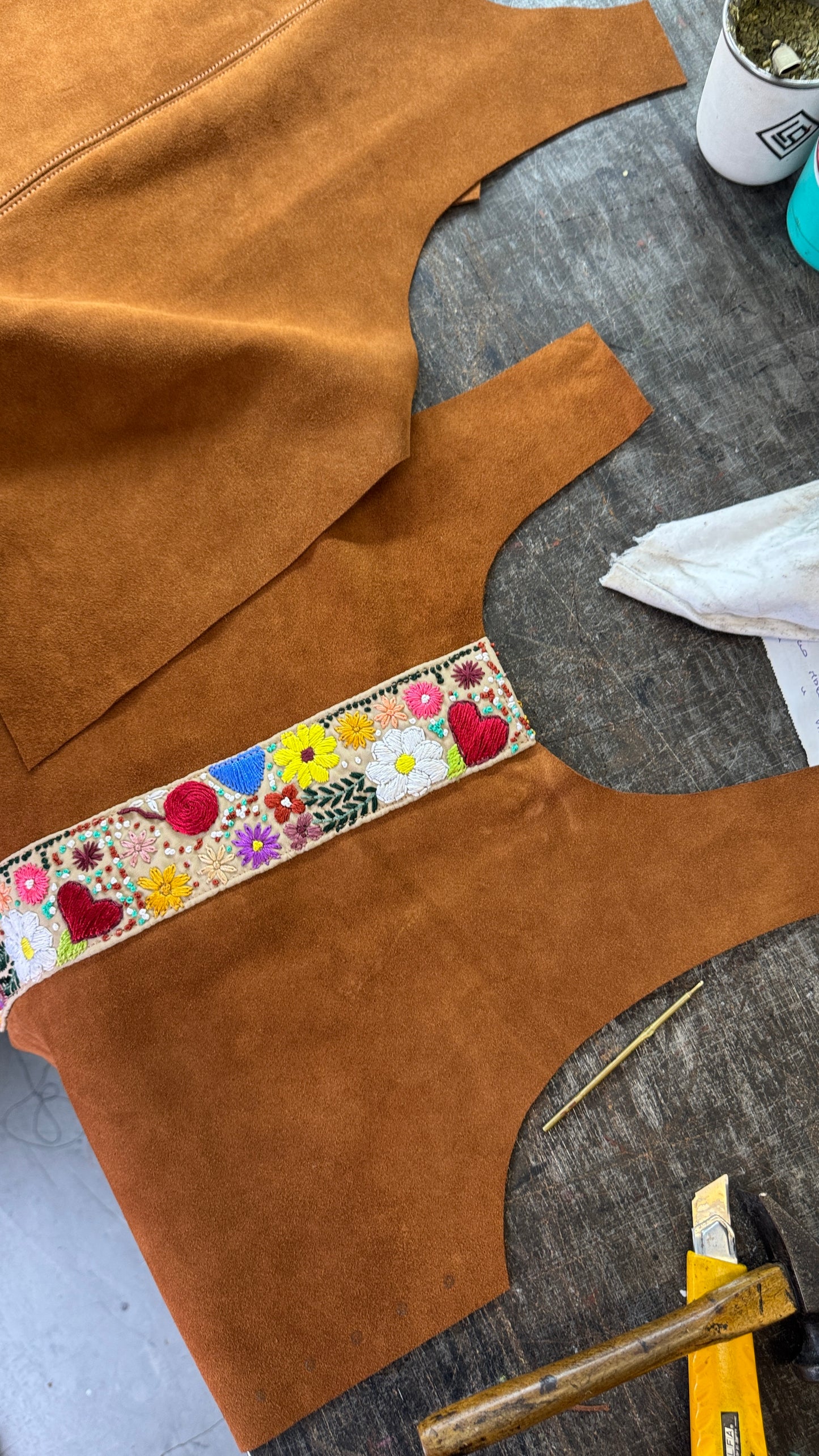 A close-up of a brown suede garment or textile piece laid on a gray work surface, featuring a colorful floral embroidered band across the chest area. Other matching brown suede pieces and sewing/craft tools are partially visible around it.