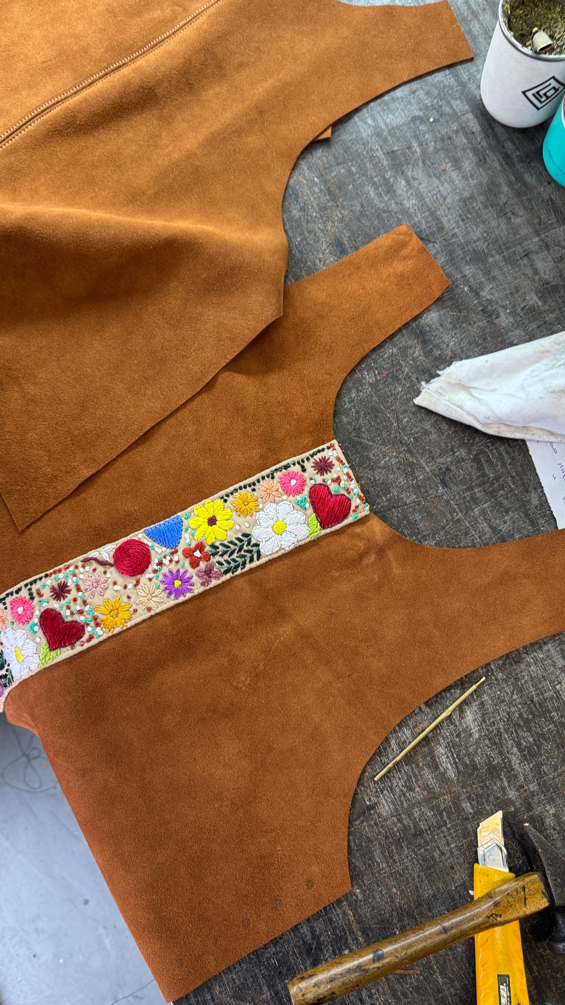 A close-up of a brown suede garment or textile piece laid on a gray work surface, featuring a colorful floral embroidered band across the chest area. Other matching brown suede pieces and sewing/craft tools are partially visible around it.