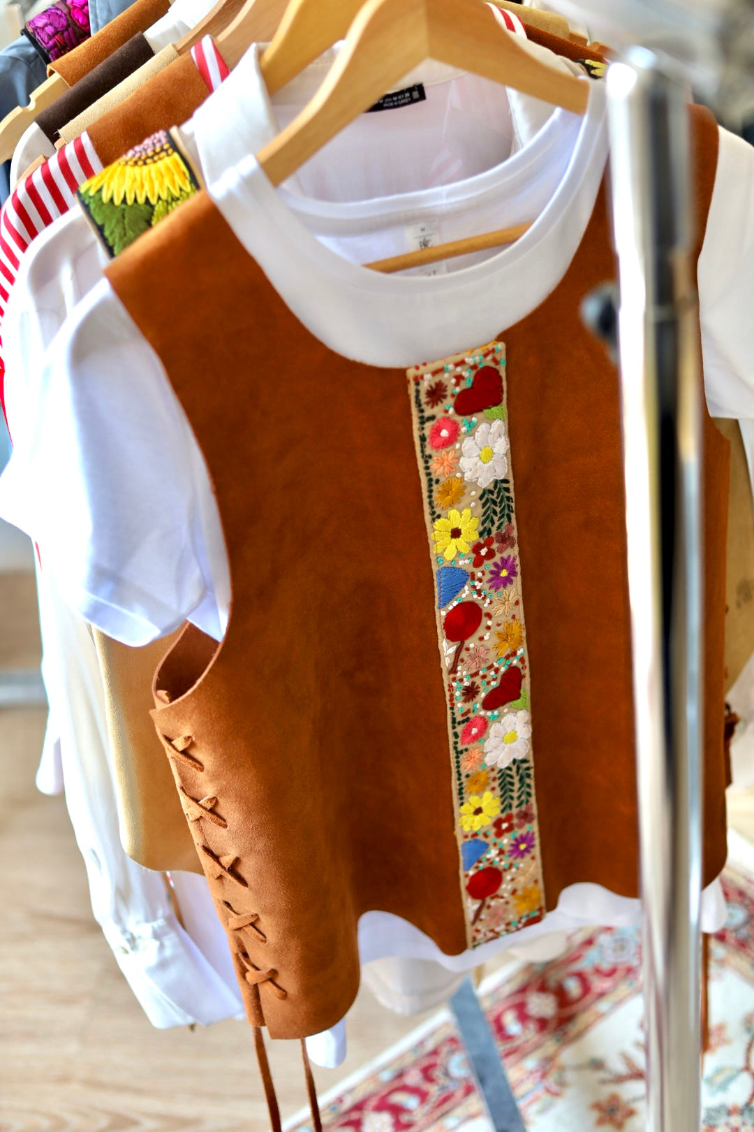 A brown suede-like sleeveless vest with a colorful floral embroidered vertical panel on the front, layered over a white short-sleeve top on a clothing rack.