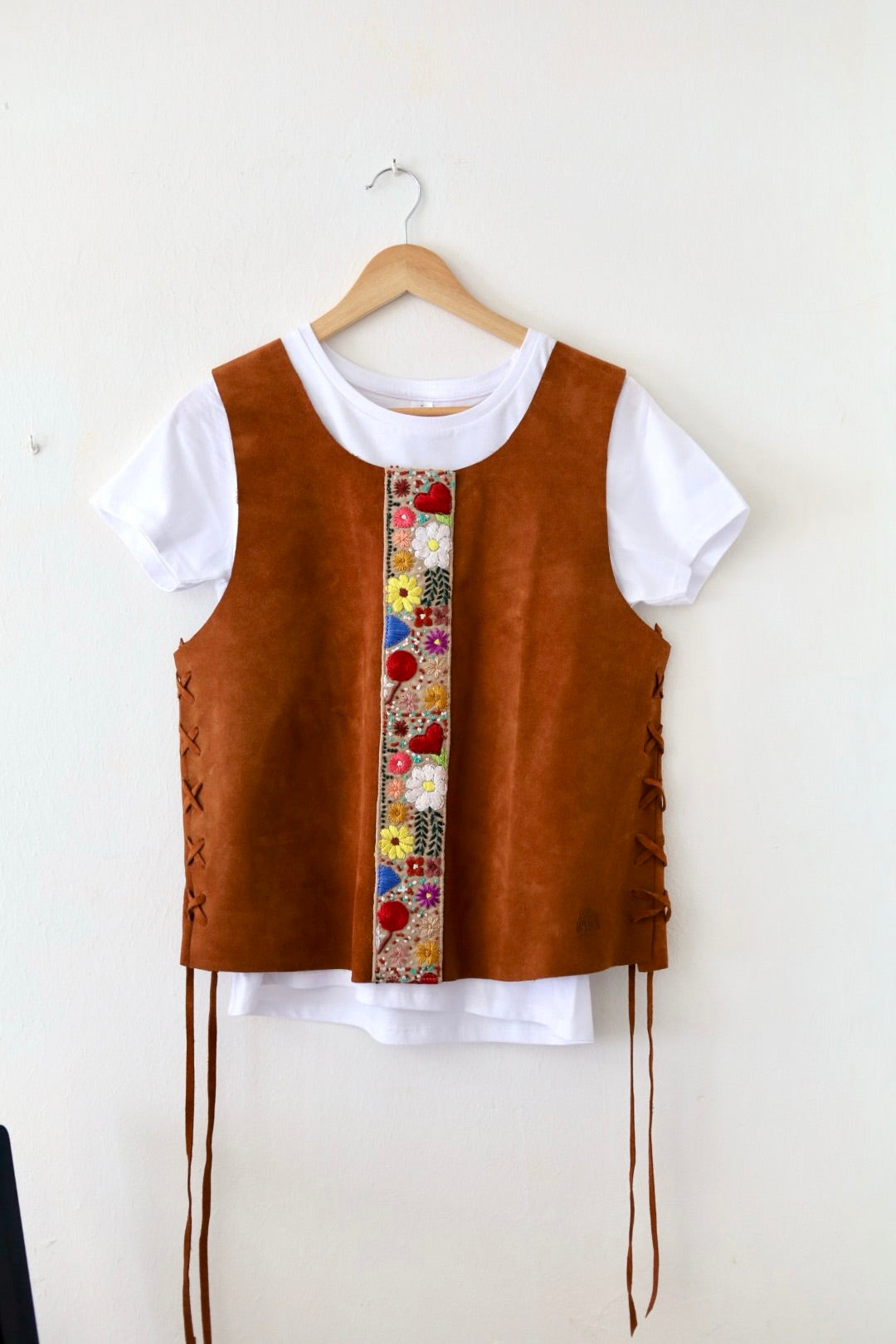 A brown suede-like vest layered over a white short-sleeve t-shirt, hanging on a wooden hanger against a plain off-white wall. The vest features a vertical decorative panel with colorful floral embroidery down the center and lace-up detailing on the sides.