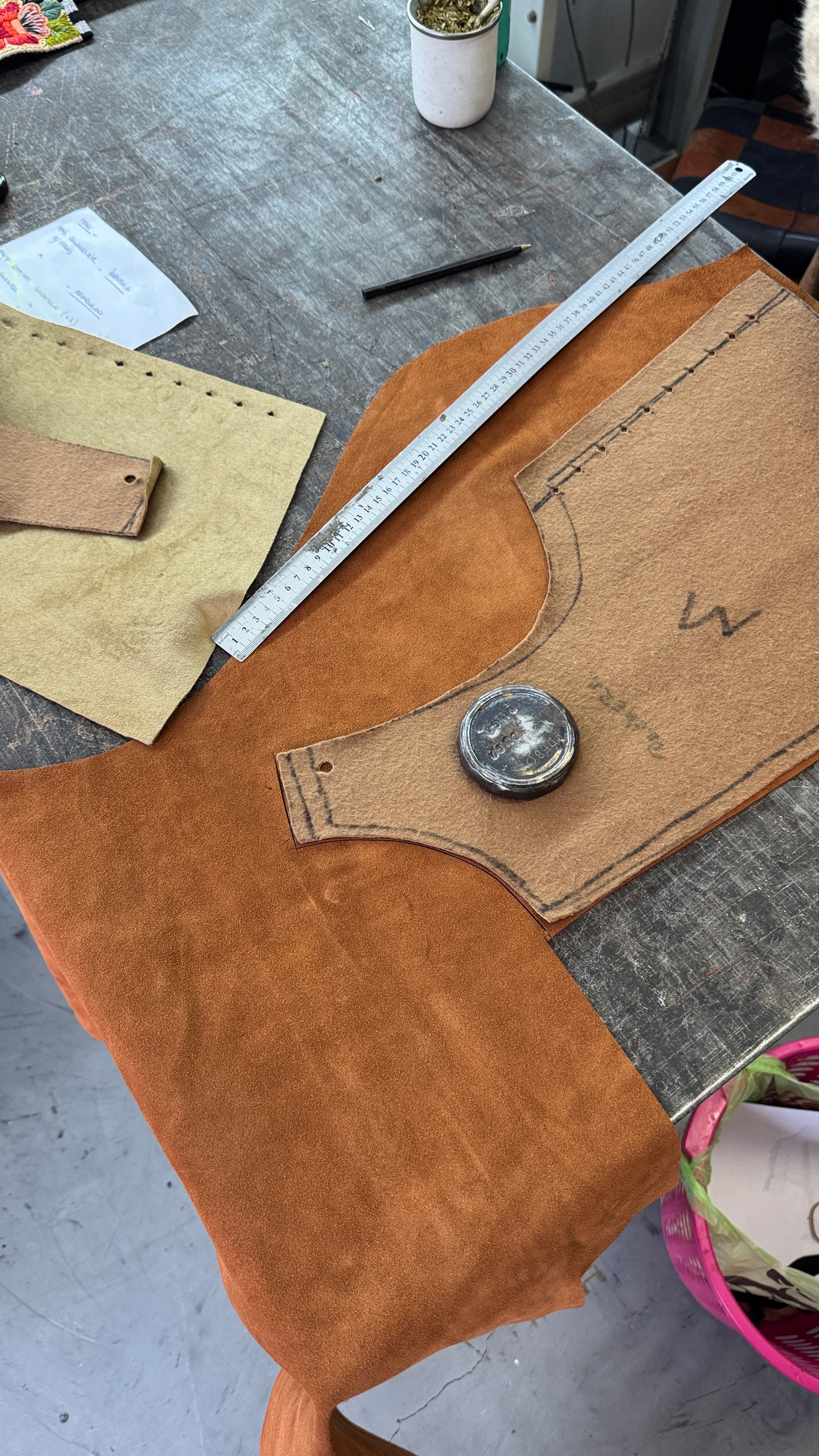 A leathercraft or tailoring workspace showing a piece of orange-brown suede or leather fabric laid on a gray workbench, with a tan paper pattern template, metal ruler, and small tools nearby.