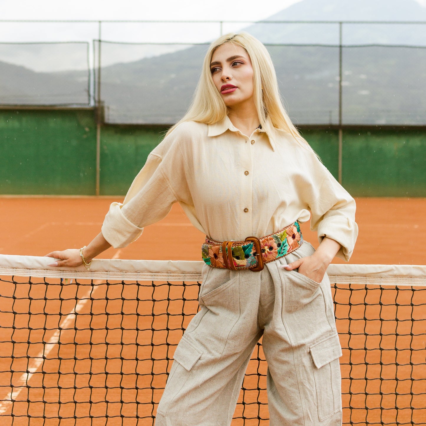 Woman standing on a tennis court wearing a beige outfit with a colorful belt.