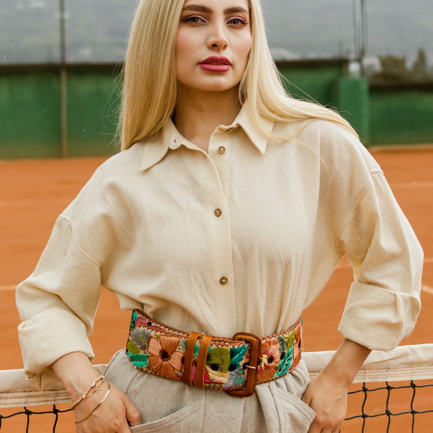 Woman standing on a tennis court wearing a beige outfit with a colorful belt.