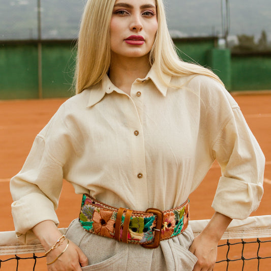 Woman standing on a tennis court wearing a beige outfit with a colorful belt.