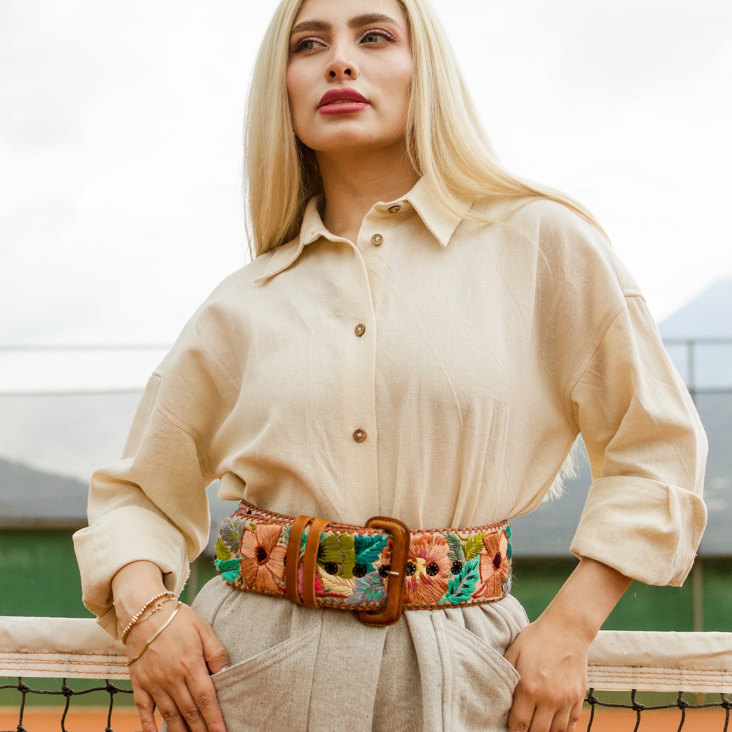 Woman wearing a beige shirt and pants with a colorful belt, standing on a tennis court.