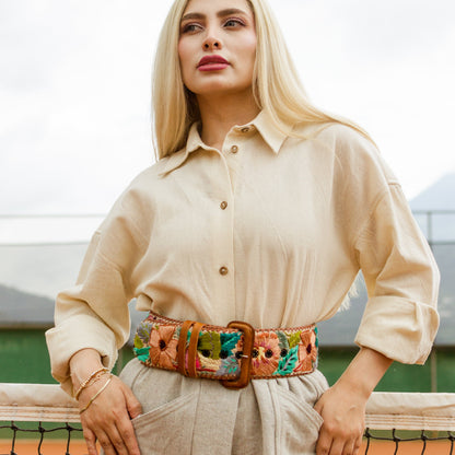 Woman wearing a beige shirt and pants with a colorful belt, standing on a tennis court.