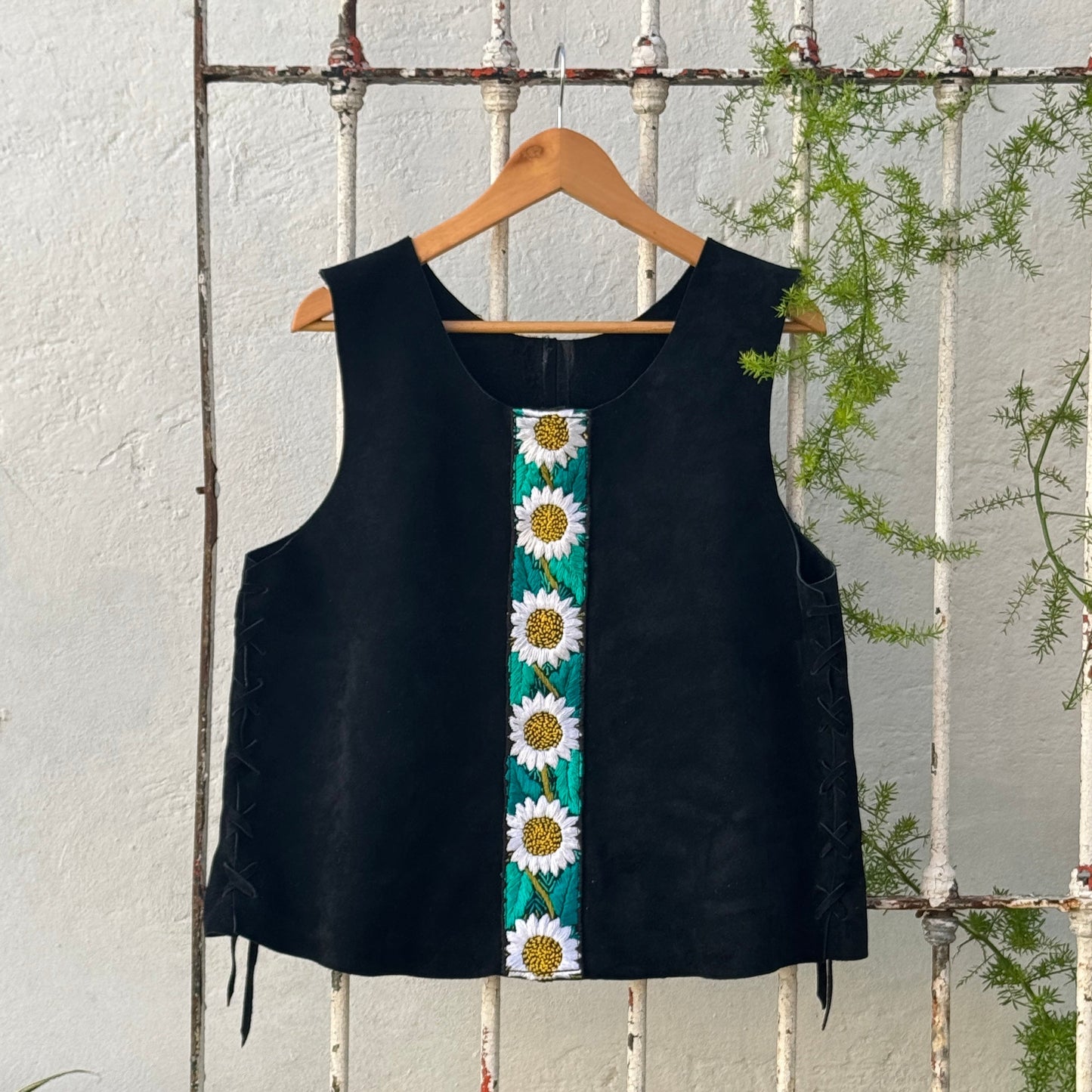 Black sleeveless top with sunflower pattern on a wooden hanger against a white wooden fence.