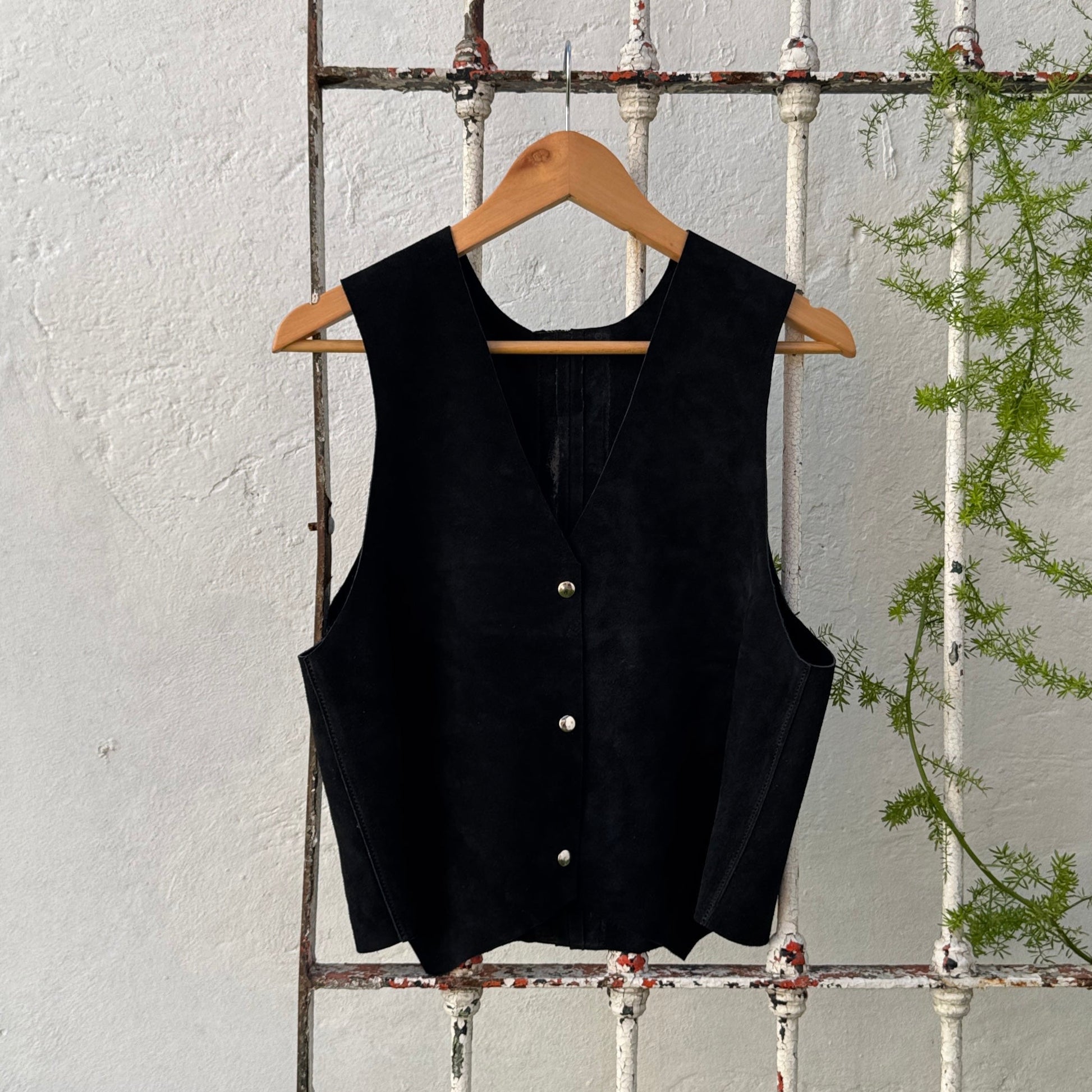 Black sleeveless top on a hanger against a white wooden panel background with greenery.