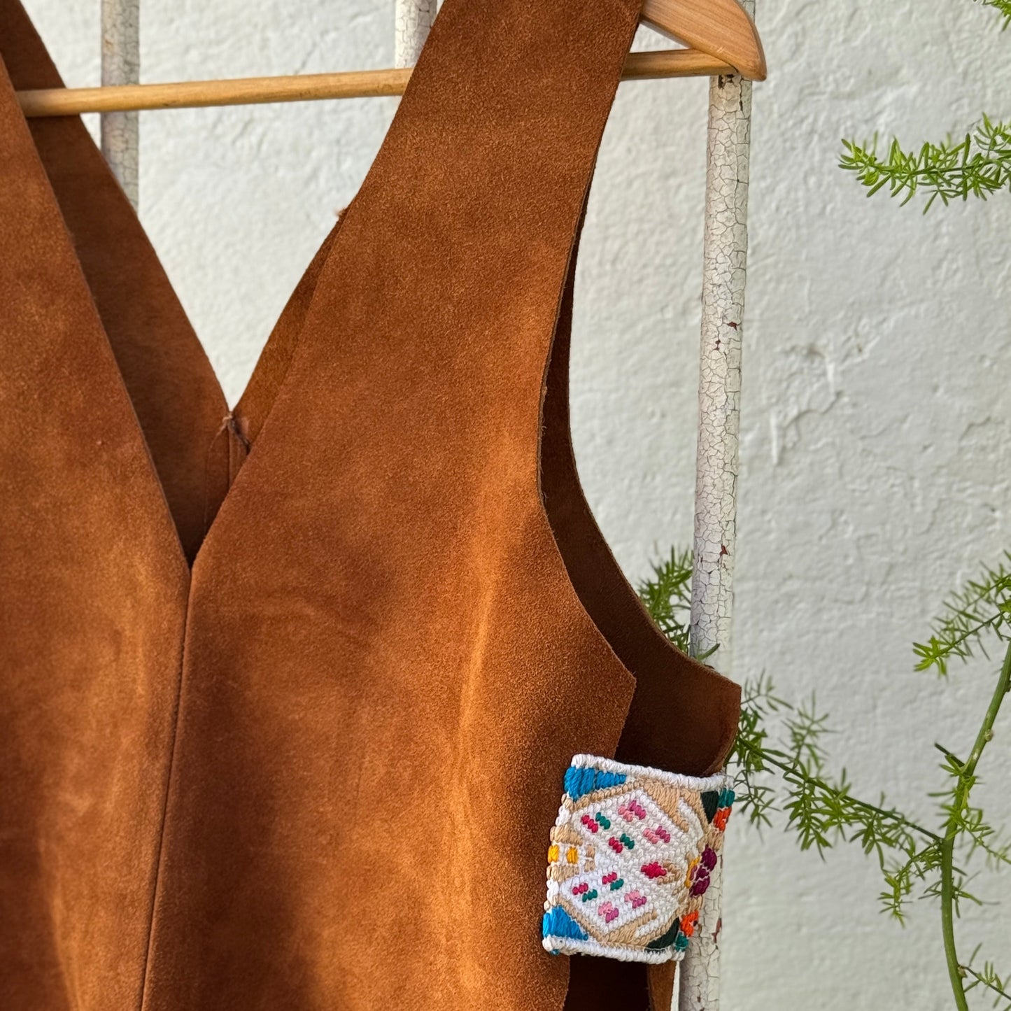 Brown suede top with a colorful patch on a hanger against a white wall.
