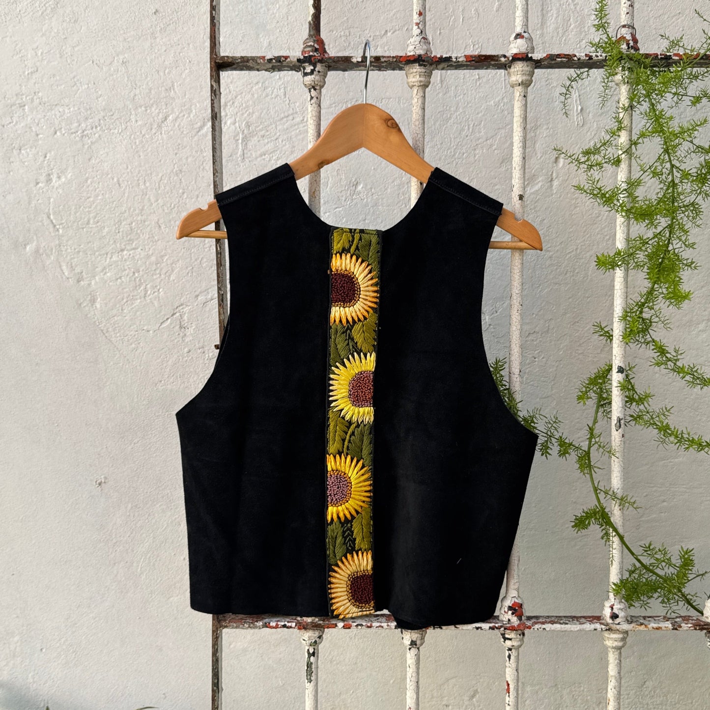 Black sleeveless top with sunflower pattern on a wooden hanger against a white wall with plants.