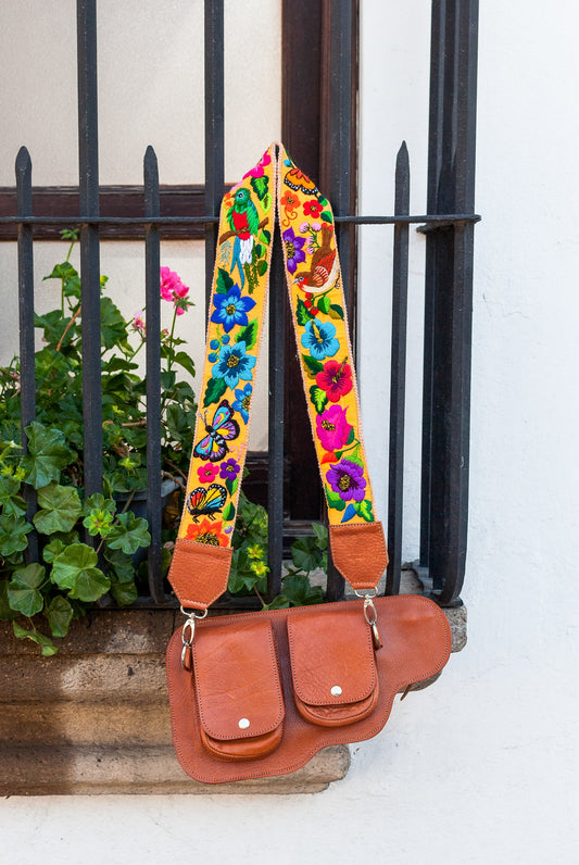 A brown leather shoulder bag with a wide, vibrant hand-embroidered floral strap, hanging from a black wrought-iron balcony railing next to a white wall and green plants.