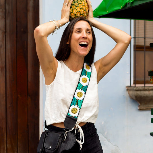 Woman holding a pineapple on her head with a colorful bag strap over her shoulder, standing in front of a building with a green awning.