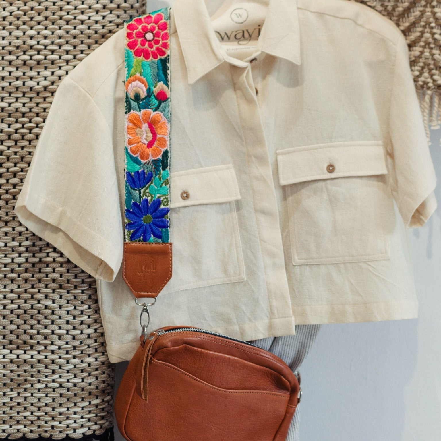 Beige jacket with floral strap and brown bag on a mannequin against a textured wall.