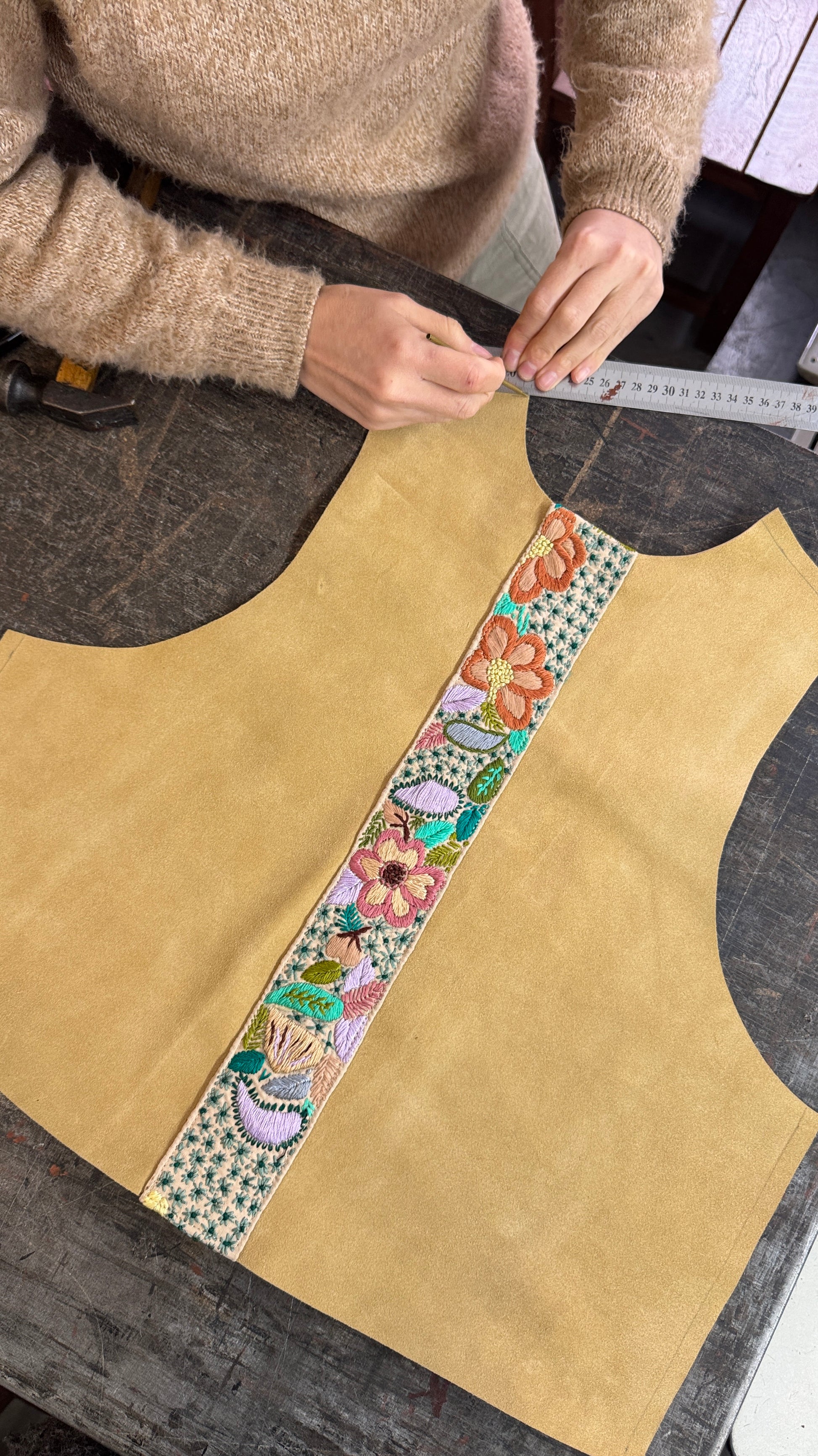 A person measures a tan suede vest with a colorful floral embroidered center stripe using a metal ruler on a dark wooden workbench.