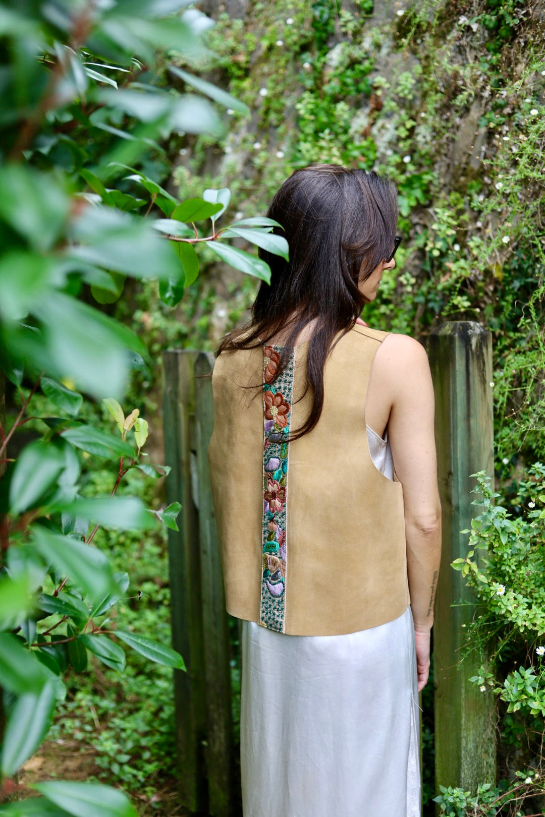 A woman viewed from behind wearing a tan sleeveless top with a colorful embroidered vertical panel down the back, paired with a white skirt, standing outdoors in front of a wooden fence and green foliage.