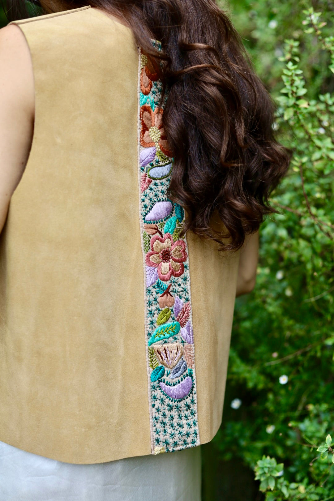 A close-up of a person from behind wearing a sleeveless tan vest with a wide, colorful embroidered vertical panel down the back, set against a background of green foliage.